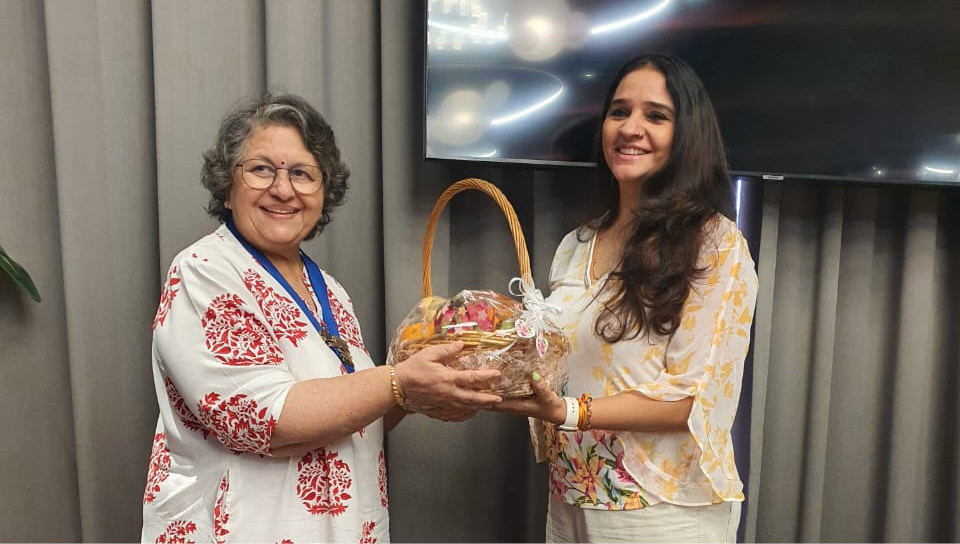 Two adults smiling and standing indoors near a table while one hands a basket to the other, both dressed in patterned clothing.
Description generated by AI