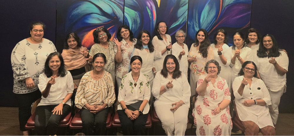 A group of people dressed mostly in white are smiling and posing together indoors in front of a large colorful abstract painting with leaf-like shapes.
Description generated by AI