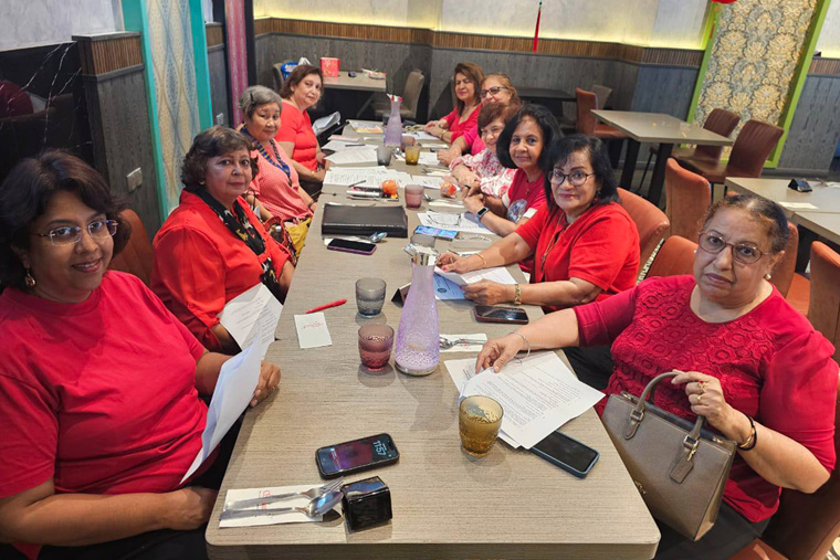 A group of people wearing red and pink clothing are seated around a long table in a restaurant, engaging with papers and electronic devices.
Description generated by AI