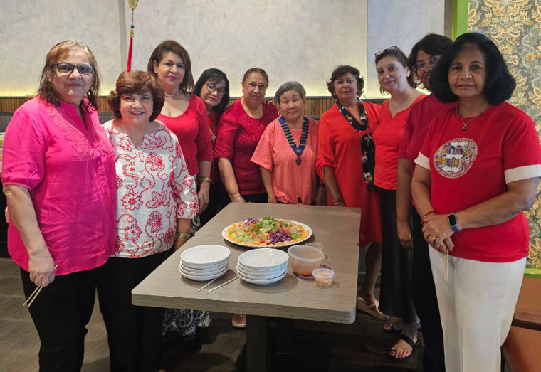 A group of adults wearing red and pink tops stand around a table with plates and a dish of food under a red hanging decoration in a warmly lit room.
Description generated by AI