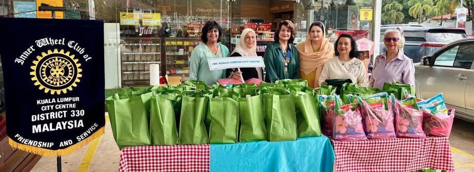 Six adults stand behind tables covered with green and colorful bags outside a Shell Select store with a banner for Inner Wheel Club of Kuala Lumpur City Centre District 330 Malaysia nearby.
Description generated by AI