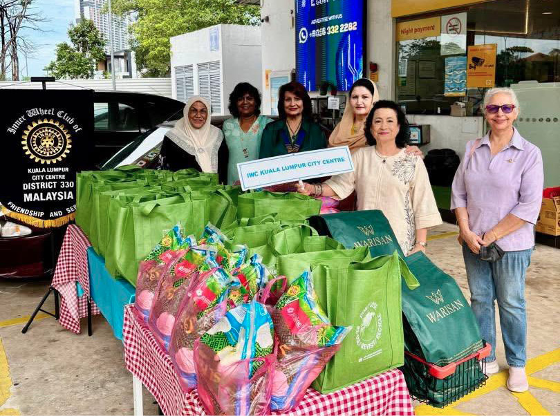 People standing behind a table covered with a red and white checkered cloth displaying green bags and colorful wrapped bouquets at an outdoor event near a gas station.
Description generated by AI