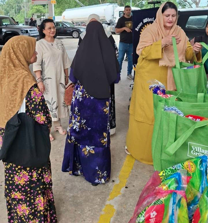 People wearing traditional clothing are gathered at a gas station near a table with green reusable bags filled with various items.
Description generated by AI
