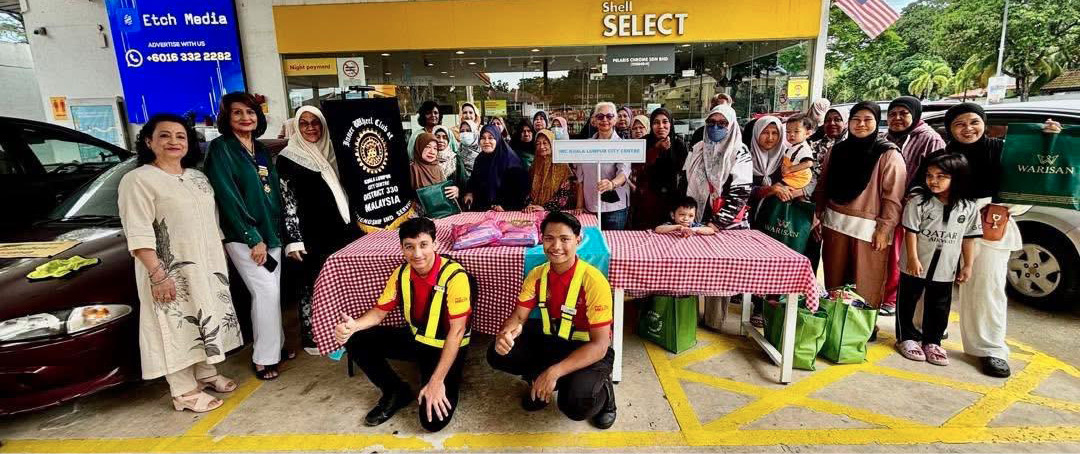 A large group of people, including two adults in yellow and red uniforms kneeling in front, are gathered around a table with a red and white checkered tablecloth outside a Shell Select store.
Description generated by AI