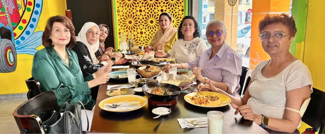 A group of seven people sitting around a table in a colorful restaurant enjoying a meal with various dishes and drinks.
Description generated by AI