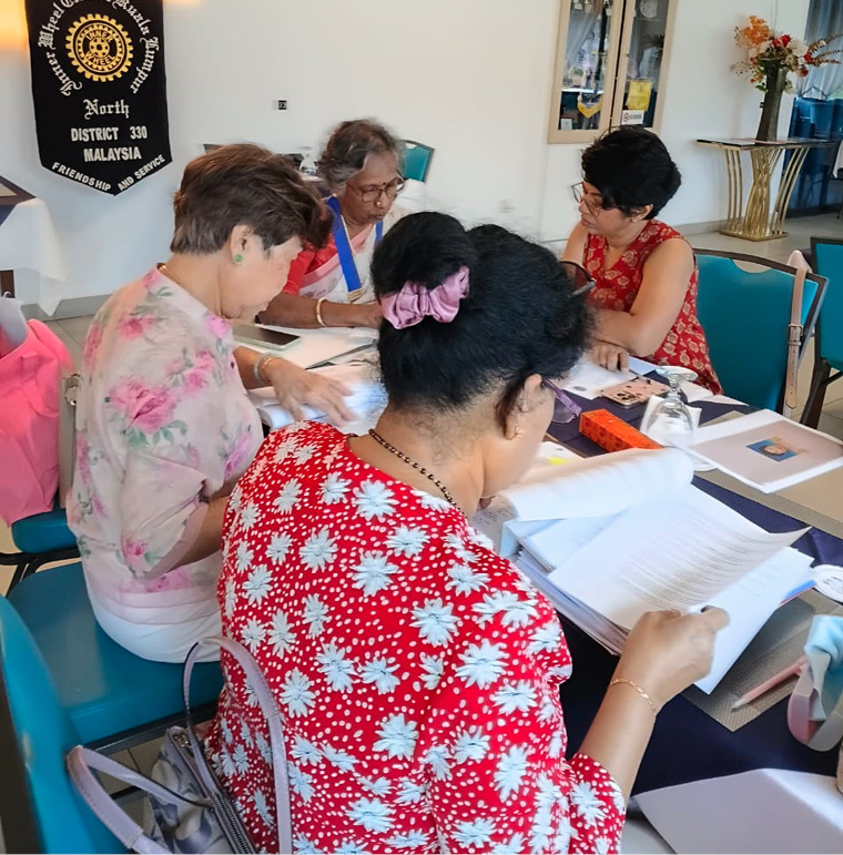 Four adults with short hair are seated around a table covered with papers and documents, engaged in discussion in a well-lit room with a banner and display cabinets in the background.
Description generated by AI