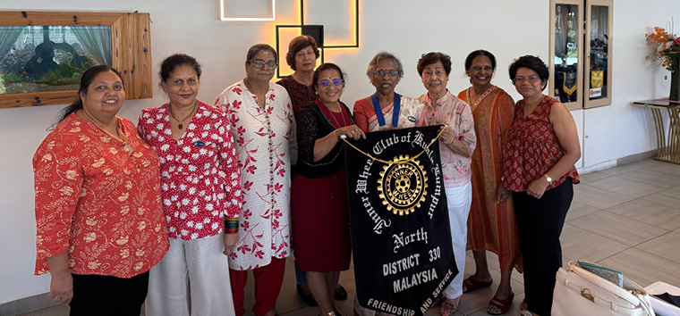 A group of eight adults wearing colorful traditional and modern clothing stand indoors, with one person holding a black banner that reads "Inner Wheel Club of Ipoh, District 330 Malaysia."
Description generated by AI