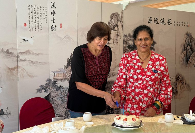 Two adults, one wearing a red floral top and the other a black and red top, are smiling and cutting a cake together at a table set with cups and plates in front of a decorative screen with calligraphy and landscape art.
Description generated by AI