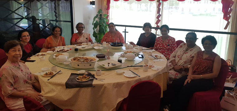 A group of people sitting around a large round table with a lazy Susan, enjoying a meal together in a well-lit room with red curtains.
Description generated by AI