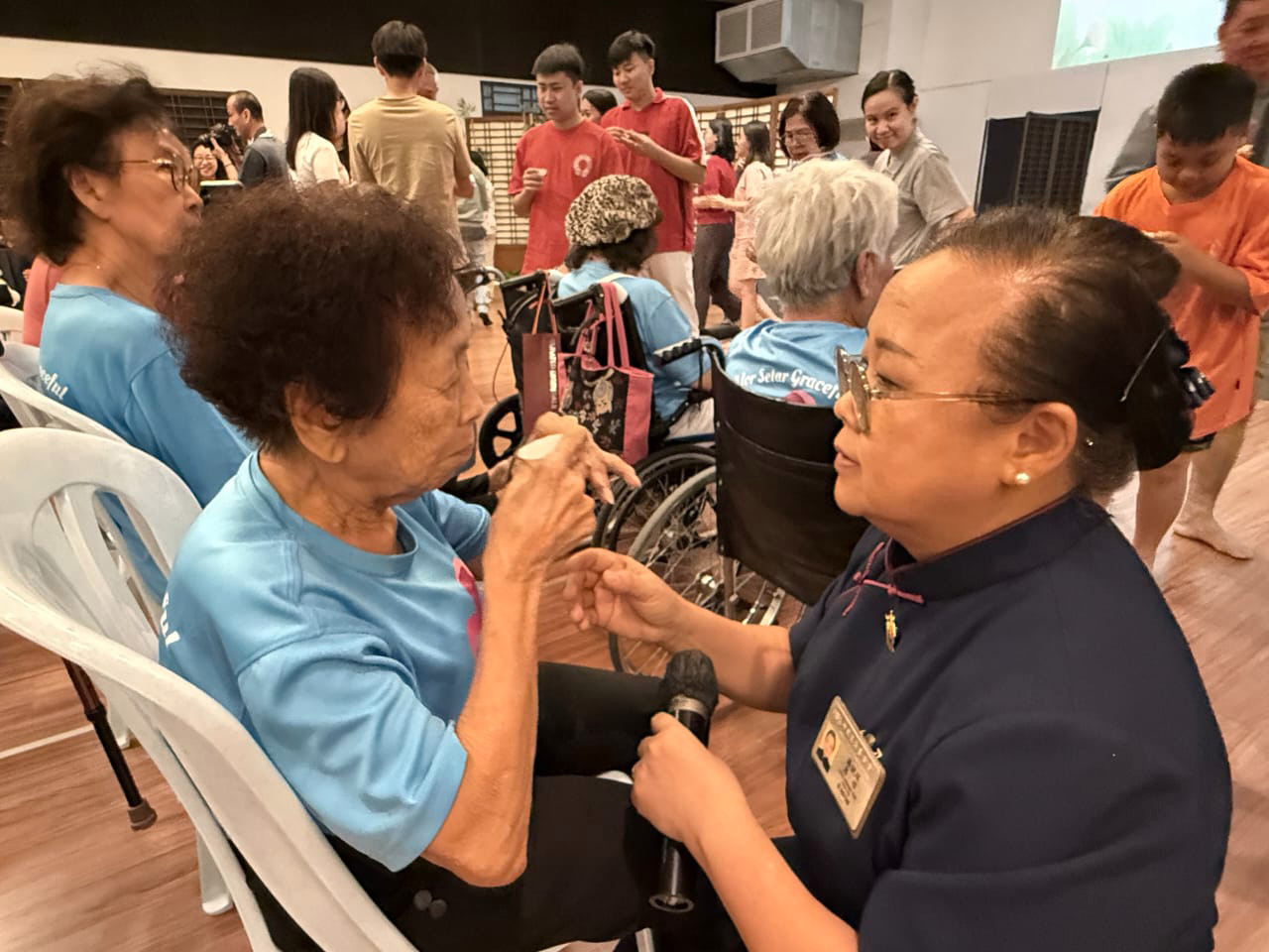 Two women are sitting in chairs, one of them is an elderly woman with a blue shirt. The other woman is a caregiver, possibly a nurse, who is assisting the elderly woman. The elderly woman is wearing a blue shirt and has a cane. The caregiver is wearing a blue shirt as well.

AI generated content