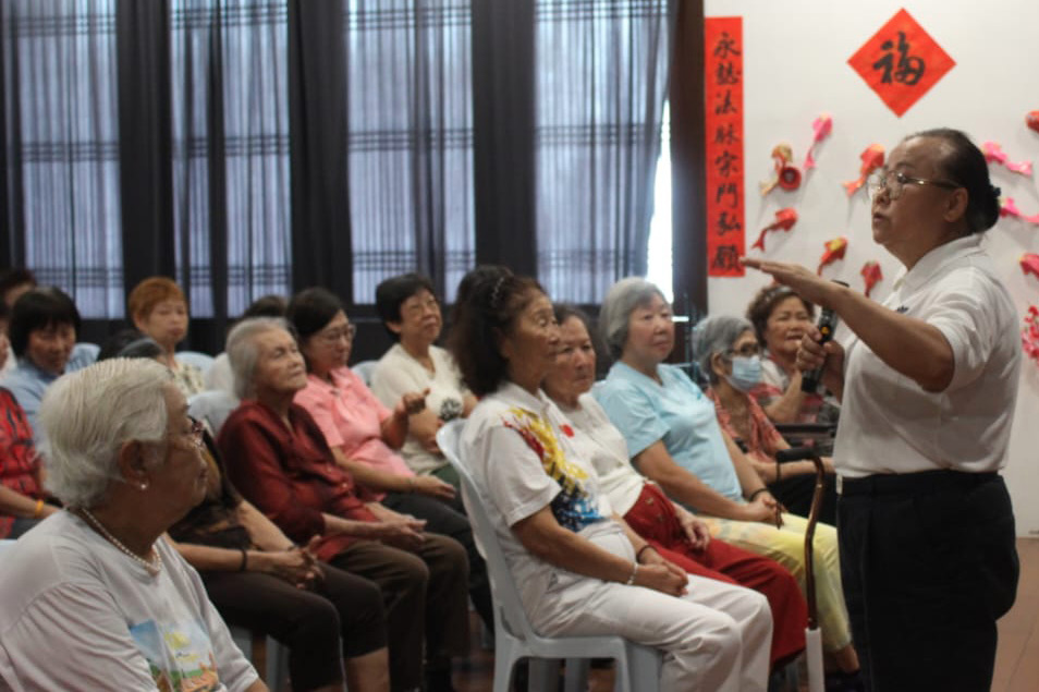 A man is giving a presentation to a group of people, who are seated in chairs. The man is wearing a white shirt and black pants. The audience is composed of several people, some of whom are wearing ties. The presentation is taking place in a room with a red and white decoration on the wall.

AI generated content