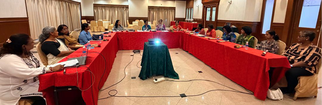 People seated around a U-shaped table covered with red cloth in a conference room with a projector in the center.
Description generated by AI