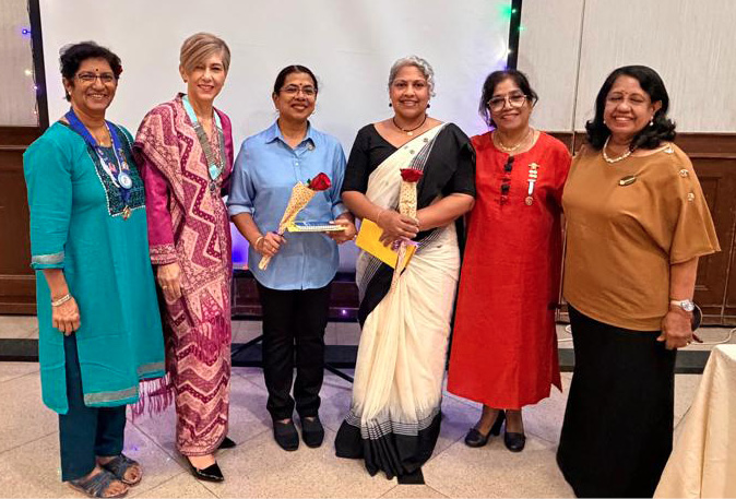Six people dressed in colorful traditional and modern clothing stand in front of a decorated backdrop with string lights and hold awards or flowers, posing for a group photo at an event.
Description generated by AI