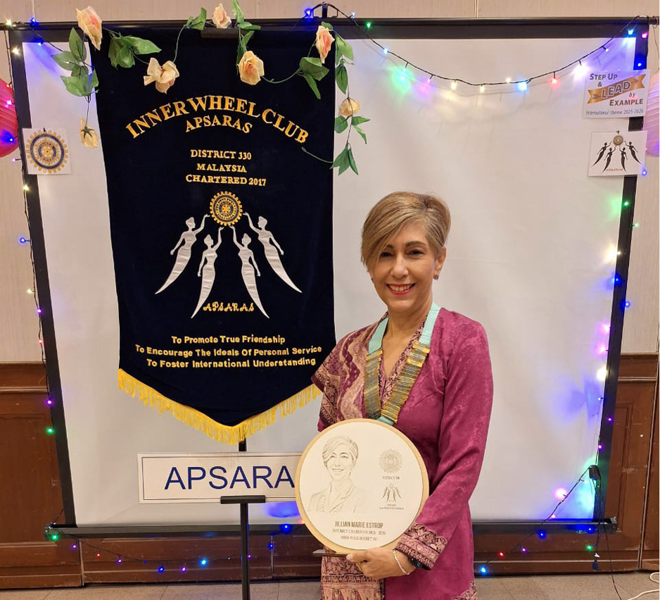 A person dressed in traditional patterned clothing stands holding a circular plaque in front of a banner and sign that read "Inner Wheel Club Apsaras."
Description generated by AI