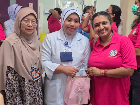 Three adults stand together in a hallway with one wearing a white medical uniform and holding a pink gift bag while the other two wear casual clothing, and several other people are visible in the background.

Description generated by AI