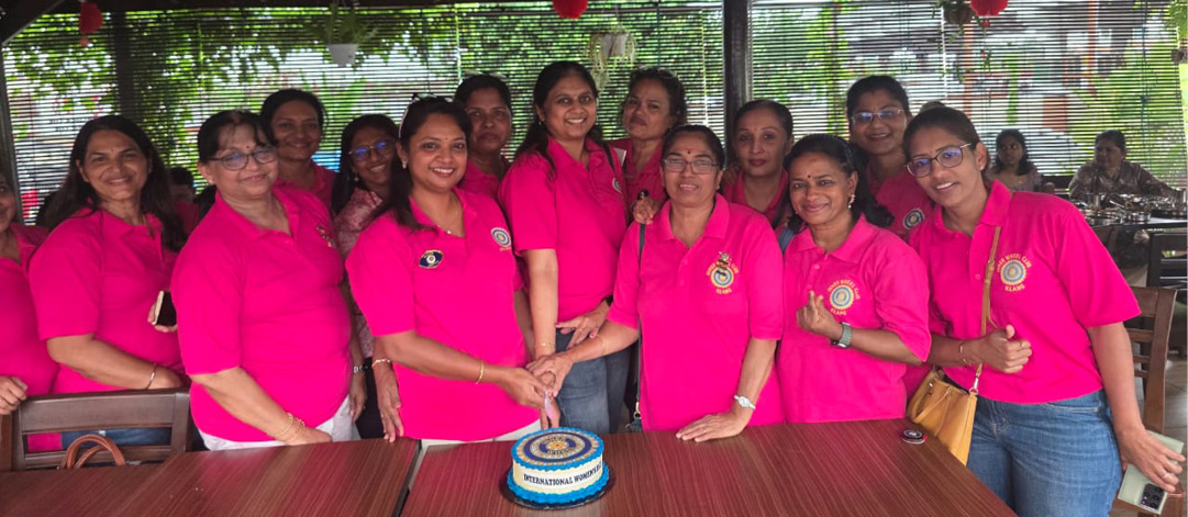A group of people wearing matching bright pink shirts are gathered around a table with a decorated cake in a casual indoor setting with wooden furniture and ceiling fans.

Description generated by AI