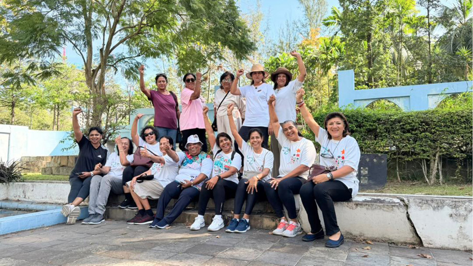 A group of people wearing casual and white event t-shirts are sitting and standing outdoors on concrete steps, smiling and raising their fists in a celebratory gesture surrounded by trees and greenery.

Description generated by AI