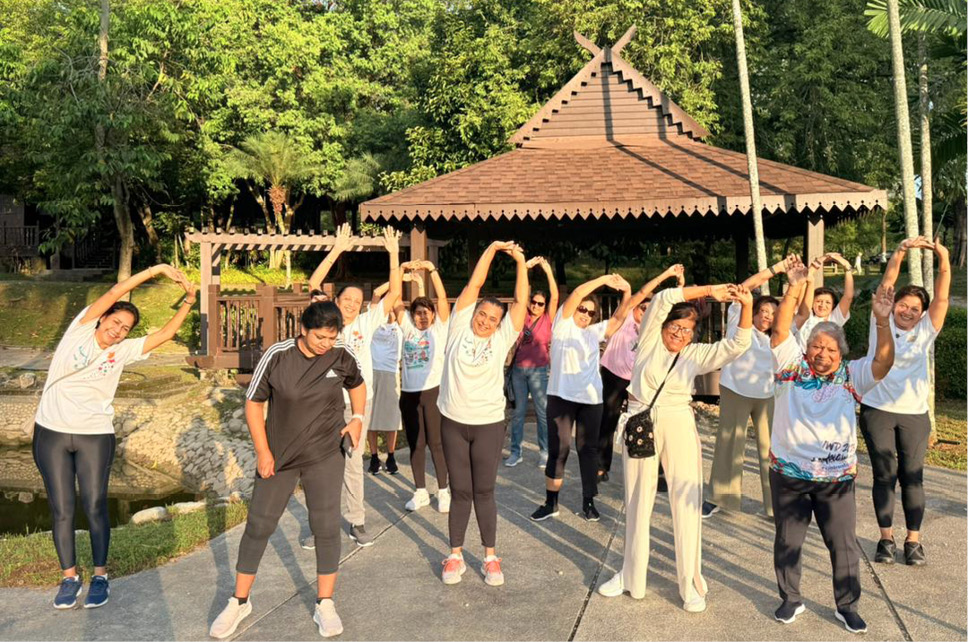 A group of people wearing casual and sportswear are standing outdoors in front of a wooden pavilion, stretching their arms above their heads in a sunny park setting.

Description generated by AI