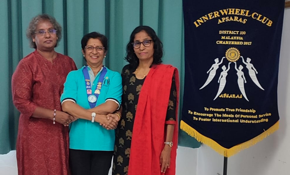 Three adults stand barefoot on a tiled floor in front of a teal curtain next to a dark blue banner with yellow text and graphics for the Inner Wheel Club.
Description generated by AI