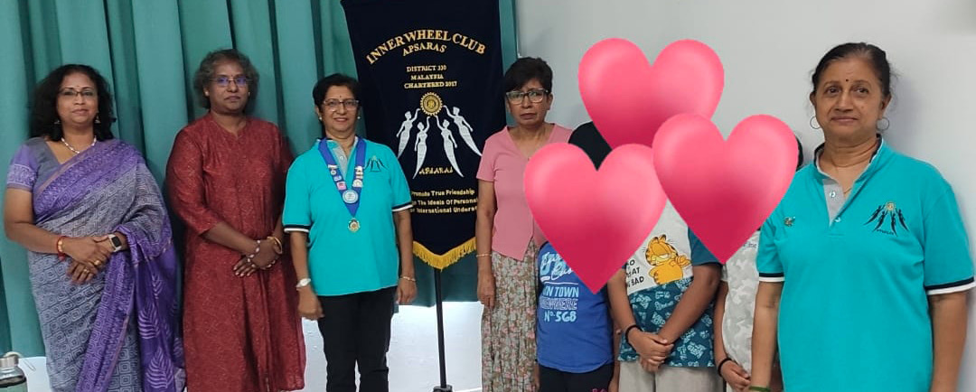 A group of seven people standing indoors near a banner with some wearing traditional clothing and others in teal shirts, with three children in front partially covered by heart emojis.
Description generated by AI