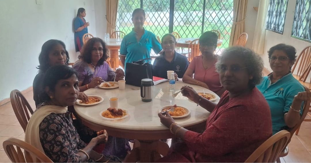 A group of people sitting around a round table eating a meal together in a bright room with large windows and curtains.
Description generated by AI