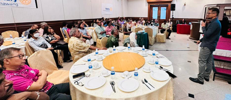 A person is giving a presentation to a seated audience in a conference room with round tables set with plates and water bottles.
Description generated by AI
