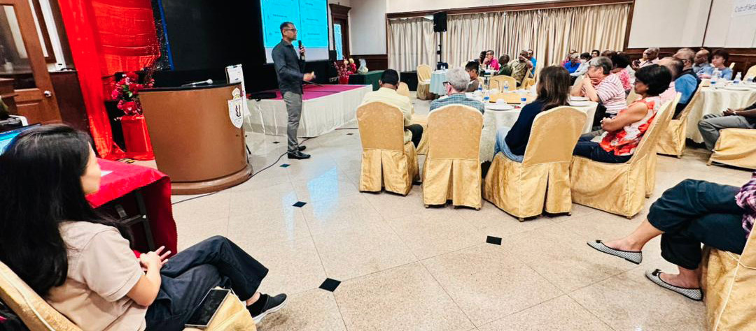 A person is giving a presentation to an audience seated at round tables in a conference room with a large screen displaying slides.
Description generated by AI