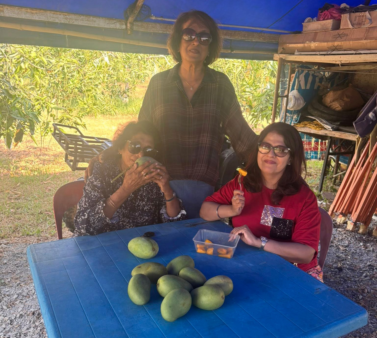 Three adults sit and stand around a blue table under a canopy with several mangoes and a container of sliced fruit while one person holds a piece of fruit on a fork.
Description generated by AI