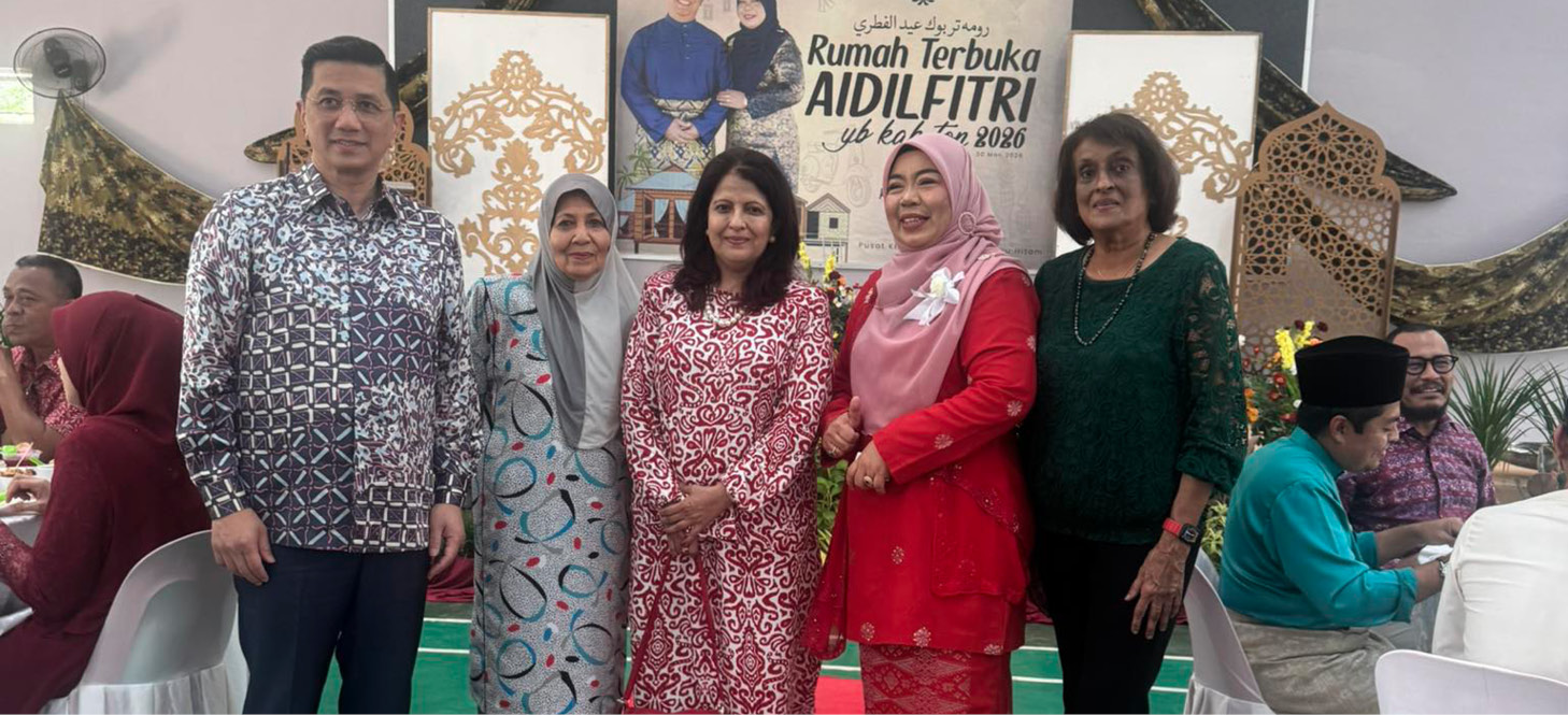 Five adults dressed in traditional festive clothing stand on a red carpet in a decorated indoor venue with a banner in the background celebrating Aidilfitri.
Description generated by AI