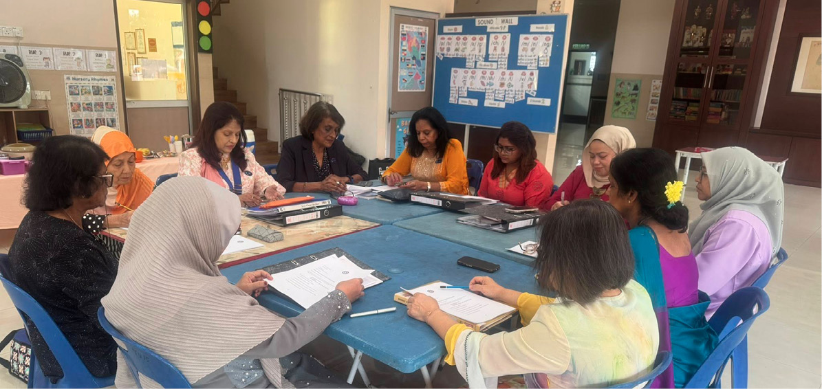 A group of adults seated around a table in a meeting room are engaged in discussion with papers and folders in front of them.
Description generated by AI