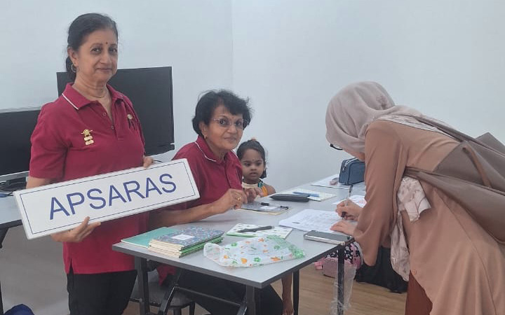 Three adults and one child are interacting at a table with papers and a sign that reads "APSARAS" in a room with wooden flooring and white walls.
Description generated by AI