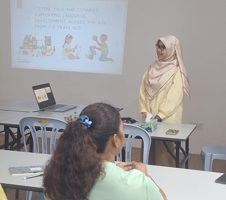 An adult wearing a light-colored headscarf and outfit is standing near a table giving a presentation to seated adults in a classroom setting with a projected slide on the wall.
Description generated by AI