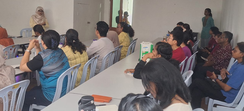A group of people seated around tables attentively listening to a person standing and speaking in a plain room.
Description generated by AI