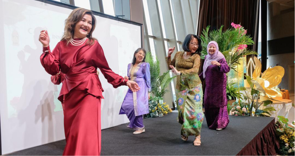 Four adults wearing colorful traditional clothing are joyfully dancing on a stage decorated with plants and flowers in a bright indoor setting.
Description generated by AI