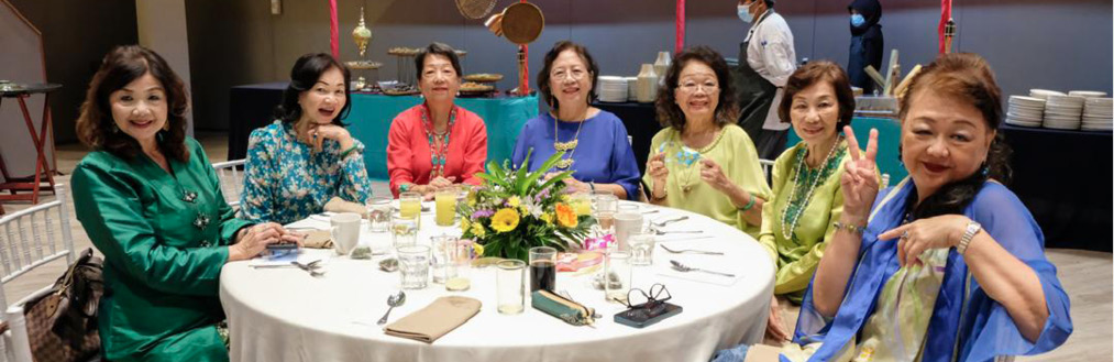 Six adults dressed in colorful clothing are seated around a round table with a white tablecloth, smiling and posing in a dining area with food stalls in the background.
Description generated by AI