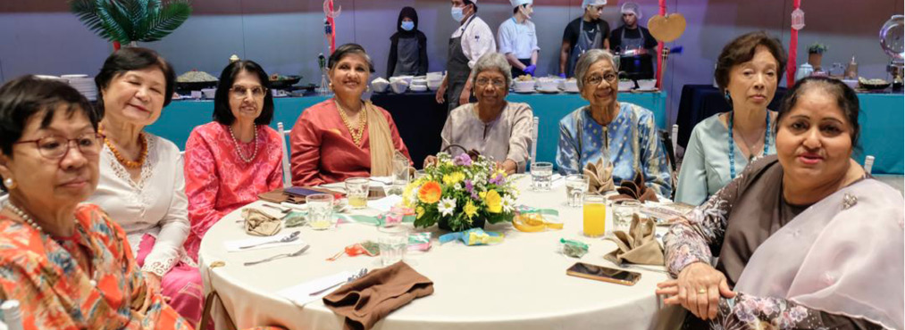 A group of people dressed in colorful traditional clothing are seated around a round table with a floral centerpiece in a banquet setting with food stalls and chefs in the background.
Description generated by AI