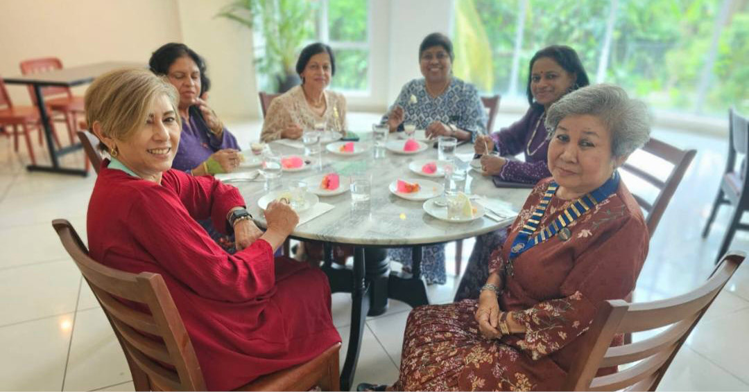 Six adults wearing colorful clothing are sitting around a round table with plates and glasses in a bright room with large windows and greenery outside.
Description generated by AI