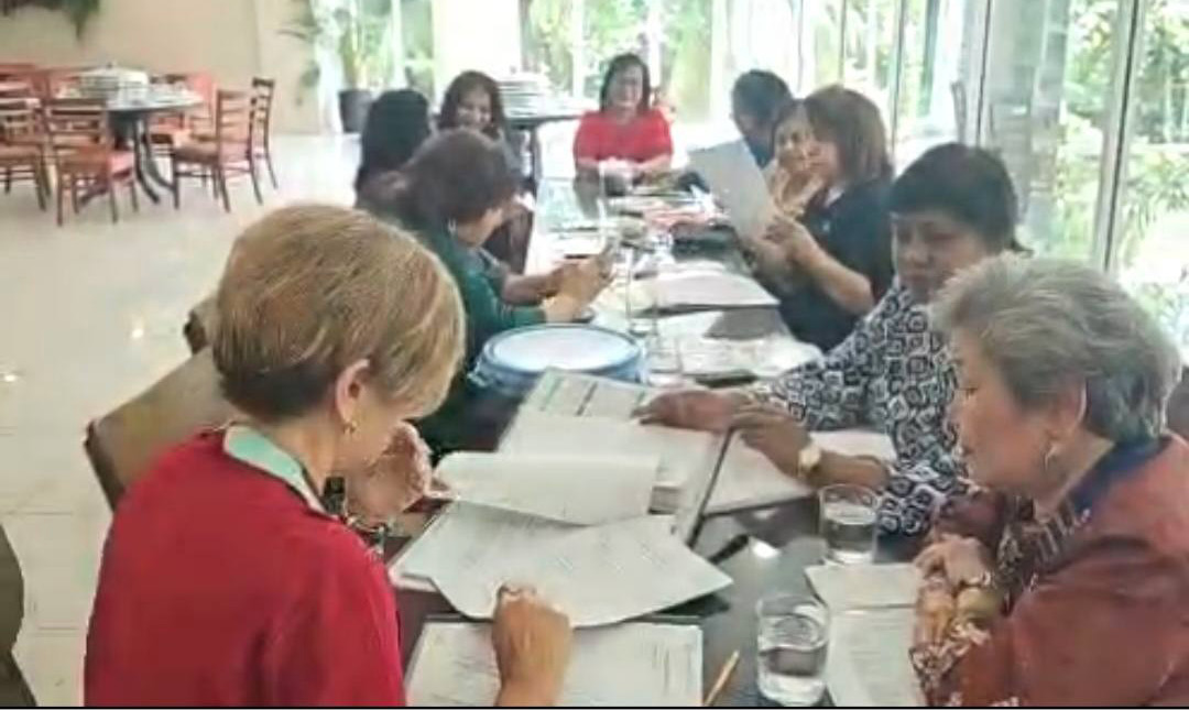 A group of adults sitting around a long table in a bright room are engaged in reading and discussing documents.
Description generated by AI