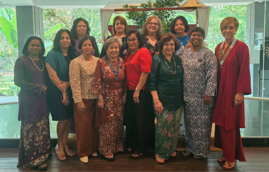 A group of adults dressed in colorful traditional and modern clothing stand closely together indoors in front of a glass wall with greenery visible outside.
Description generated by AI