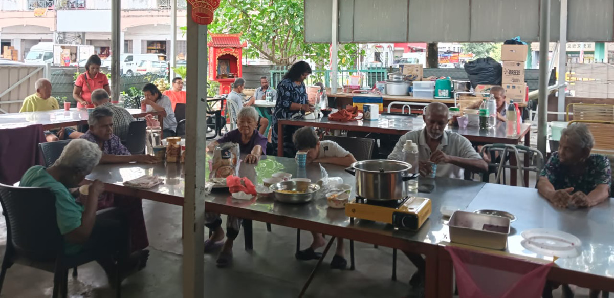 A group of adults and seniors are seated around tables in a covered outdoor area decorated with red lanterns and festive ornaments, engaging in conversation and activities with cooking equipment and food containers on the tables.
Description generated by AI