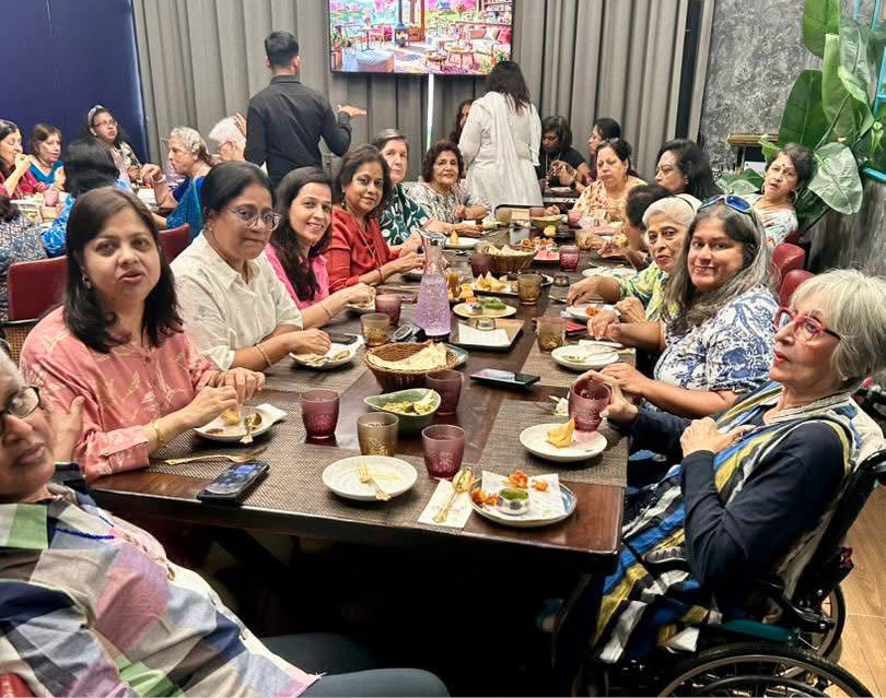 A large group of people seated around a long dining table enjoying a meal together in a well-lit room with a television and plants in the background.
Description generated by AI
