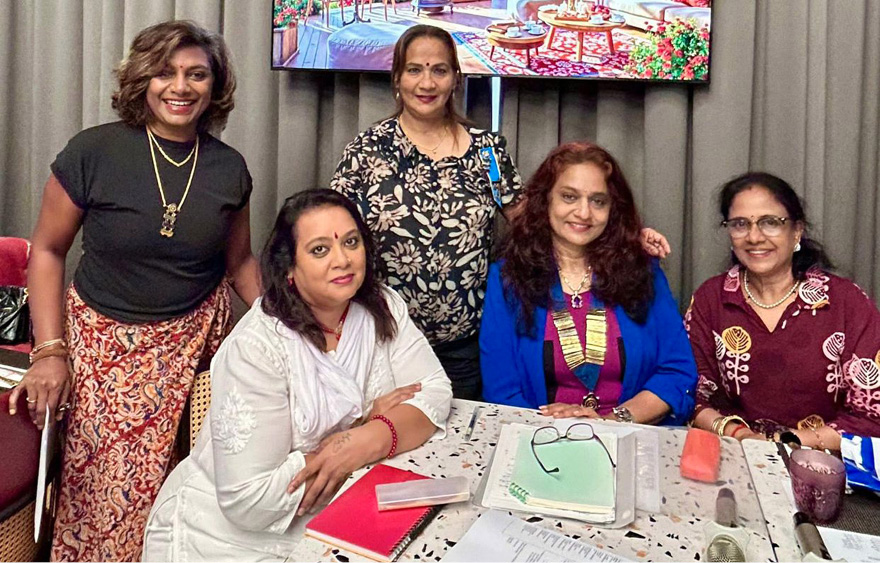 Five adults are gathered around a table with documents and notebooks, smiling and posing for the camera in a room with a large screen displaying a colorful outdoor scene.
Description generated by AI