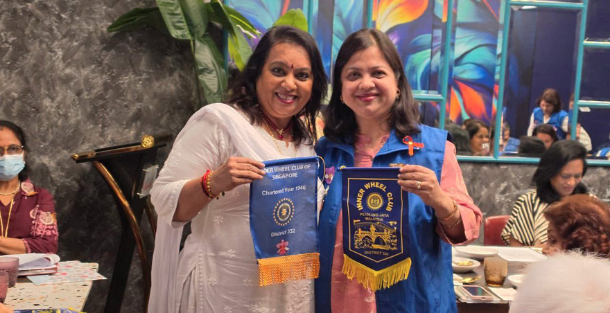 Two adults smiling and holding blue and yellow banners while standing in a room with other seated people and colorful wall art.
Description generated by AI