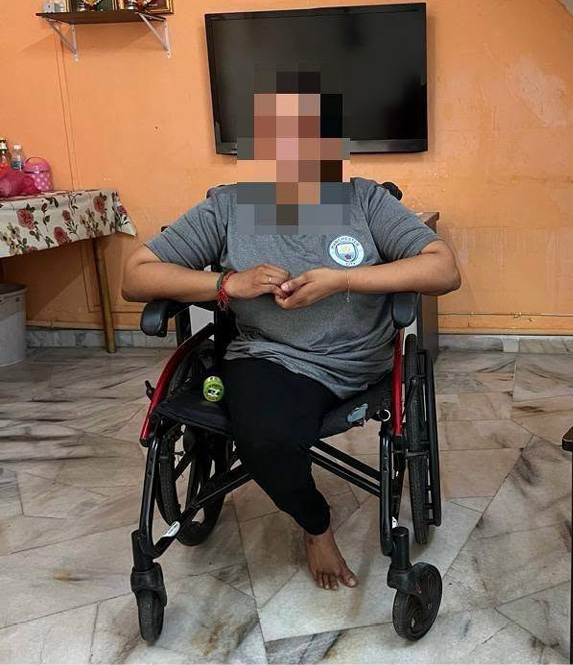 A person wearing a gray shirt and black pants is sitting barefoot in a wheelchair inside a room with an orange wall, a mounted TV, a table with a floral tablecloth, and a chair holding a white plastic bag from Allday Pharmacy.

Description generated by AI