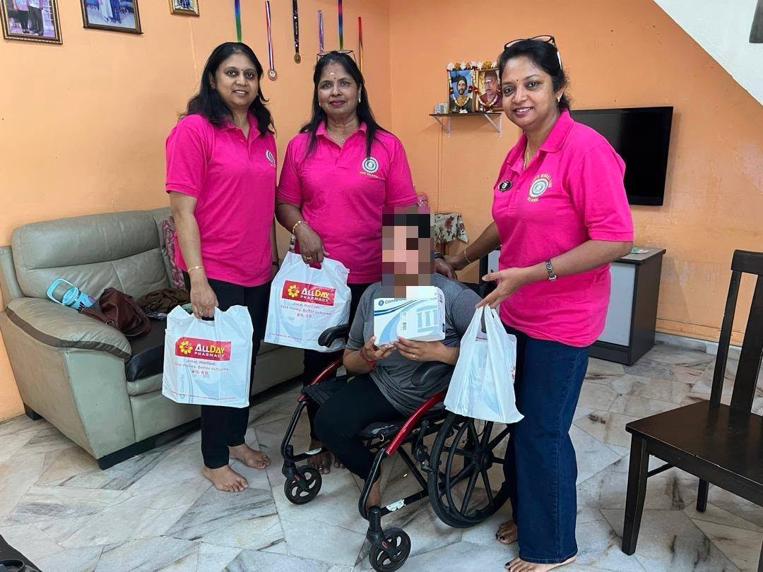 Three adults wearing matching pink shirts stand around a person in a wheelchair holding bags and a box in a living room with orange walls and marble flooring.

Description generated by AI
