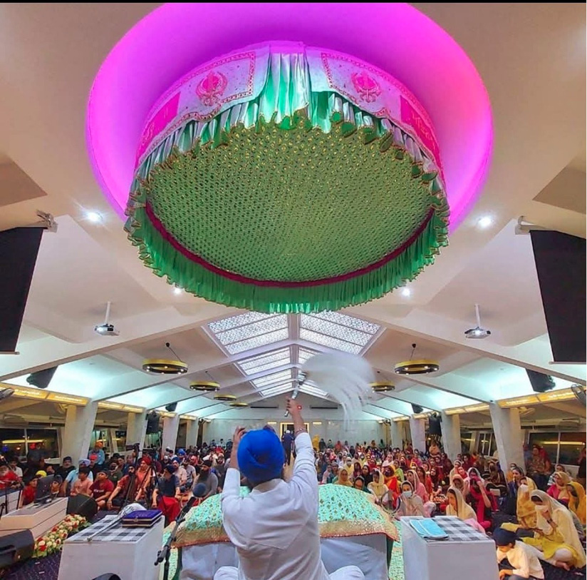 A person wearing a blue turban and white clothing raises a white cloth in front of a large crowd seated in a decorated indoor space with a large ornate canopy overhead.
Description generated by AI