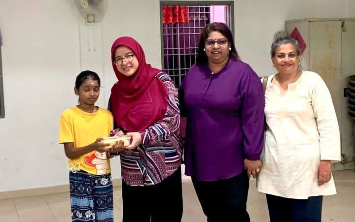 Four people stand indoors on a tiled floor with one person in a yellow shirt and patterned pants receiving an item from another person wearing a red headscarf and patterned top, while two others stand nearby smiling.
Description generated by AI
