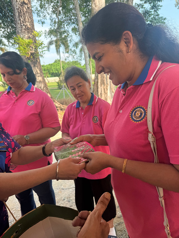 Three adults wearing matching pink shirts with blue collars are outdoors, with one person handing a decorated item to another person whose hands are visible.
Description generated by AI