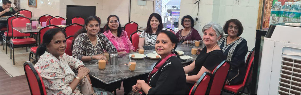 Seven adults sitting around a table in a casual dining setting with drinks and snacks, smiling and engaging with each other.
Description generated by AI