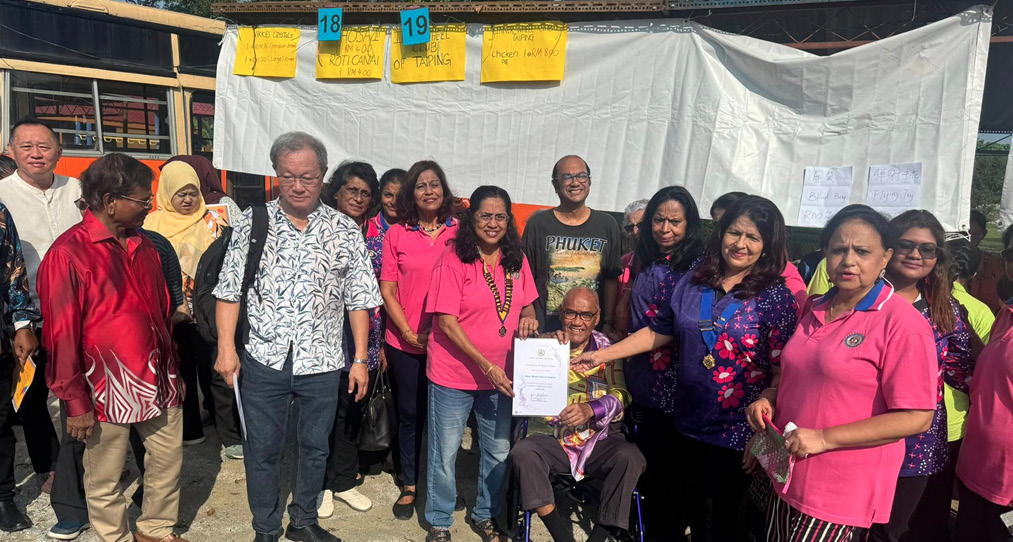 A group of people stand outdoors on gravel in front of a white backdrop with papers and yellow signs, with one person seated in a wheelchair holding a certificate while others smile and pose around them.
Description generated by AI